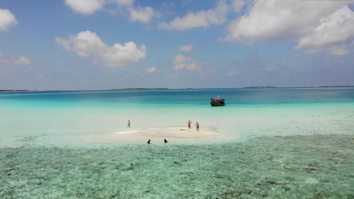 Eine Sandbank im türkisfarbenen Flachwasser, dahinter wartet ein Dhoni